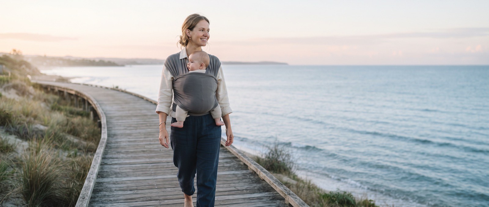 A mother with her baby in a fabric carrier walks along a wooden boardwalk beside a calm sea in the early morning.